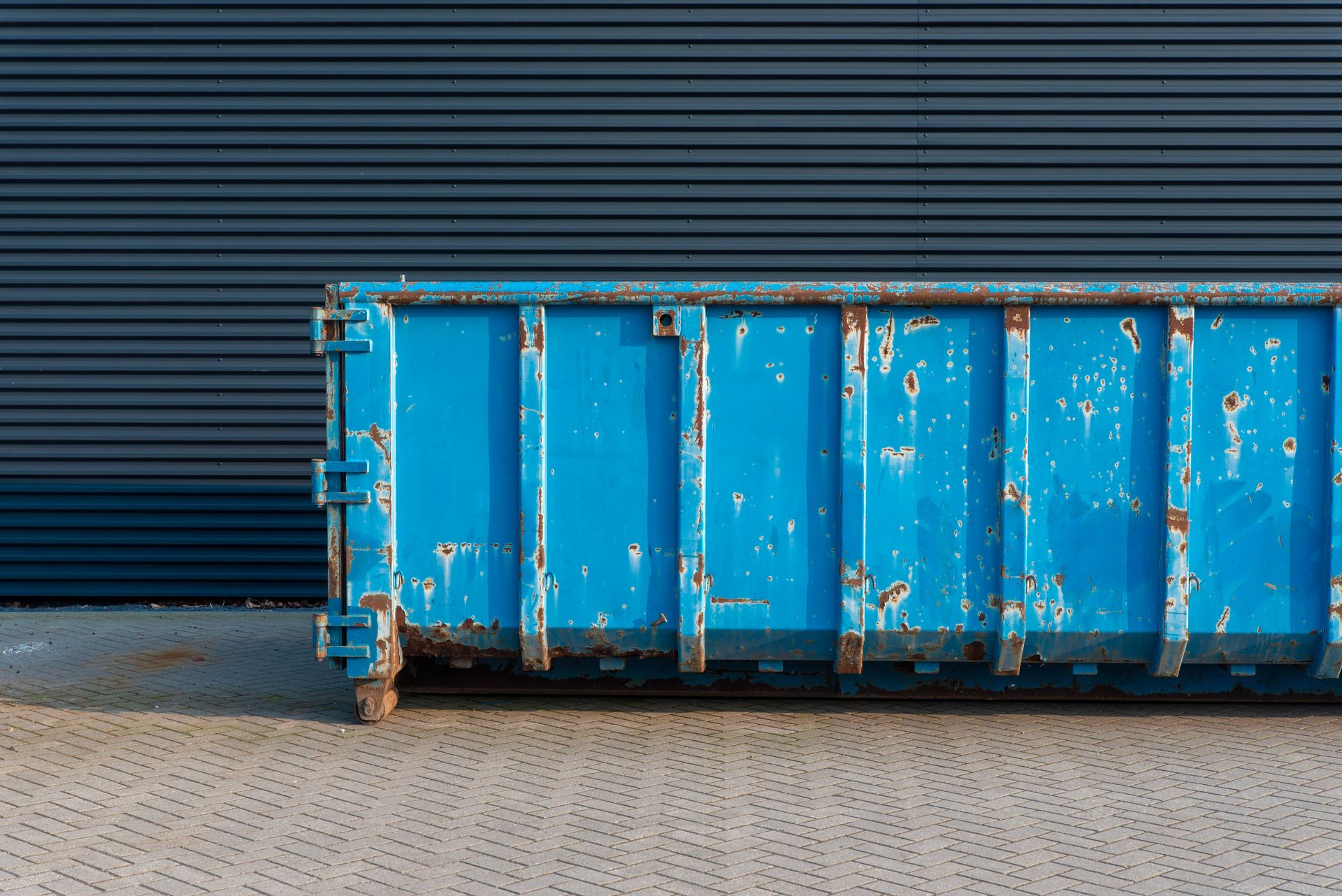 A blue rusty industrial container sits against a corrugated metal wall on a paved surface.